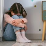 Sad little girl sitting on the floor of her bedroom with stuffed toy lying